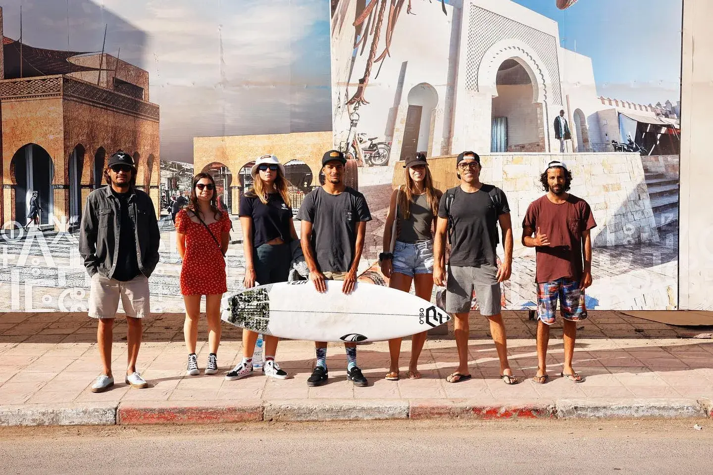 Seven people standing together with a surfboard in front of a mural of Moroccan architecture